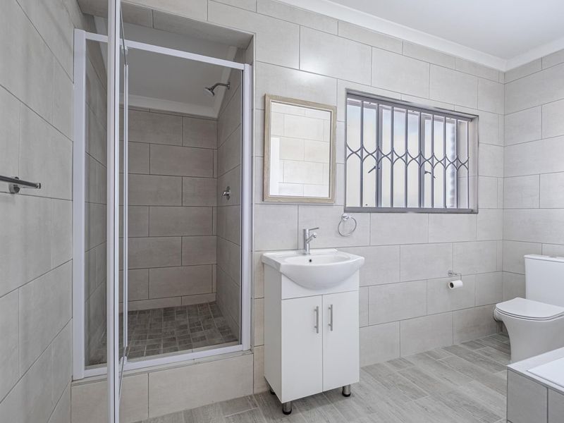 Modern bathroom featuring a walk-in shower, vanity basin, toilet, and neutral floor-to-ceiling tiling with security bars on the window.