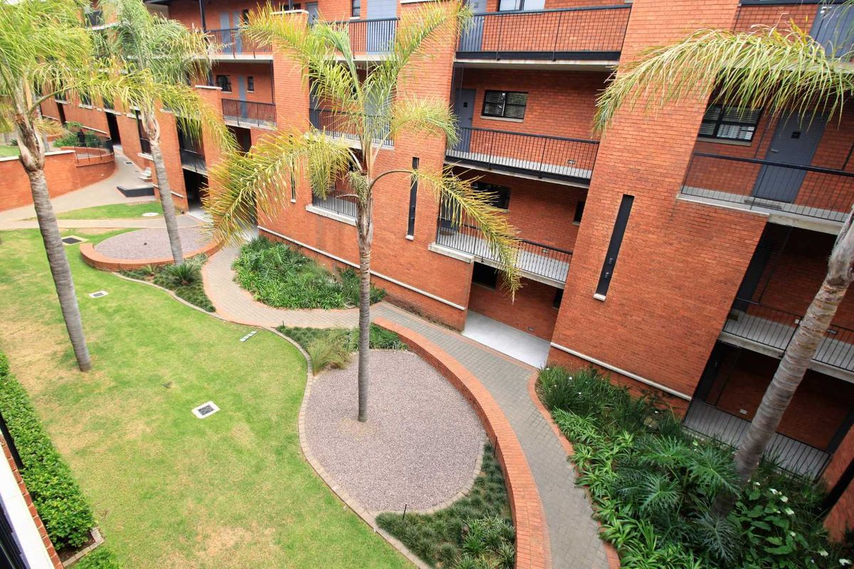 An elevated view of an apartment complex featuring a red brick facade, private balconies, and a well-maintained communal garden with paved pathways.
