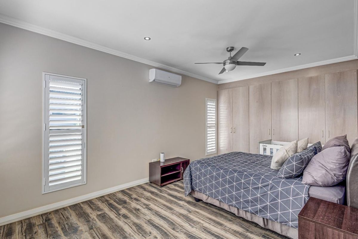 Spacious bedroom featuring built-in cupboards, plantation shutters, a ceiling fan, and a split-system air conditioner, finished with modern laminate flooring.