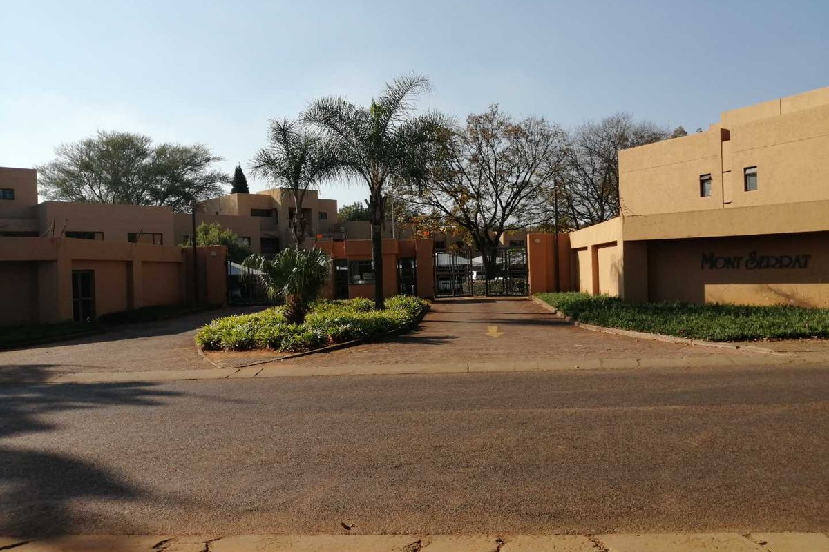 The entrance to Mont Serrat apartment complex featuring a paved driveway, secure gated access, and established landscaped garden beds.