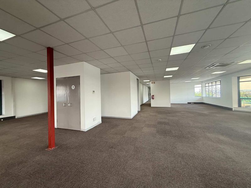 Spacious open-plan office area featuring a suspended ceiling, recessed lighting, carpeted flooring, and a central steel support column.
