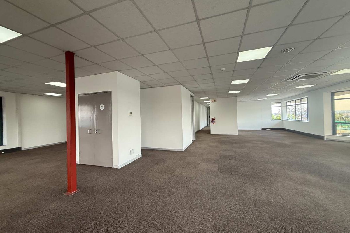 Spacious open-plan office area featuring a suspended ceiling, recessed lighting, carpeted flooring, and a central steel support column.