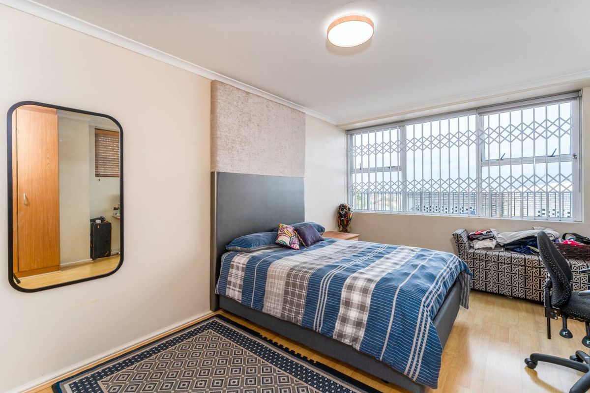 A bedroom featuring light wood-look flooring, a built-in headboard feature wall, and a large window fitted with security bars.