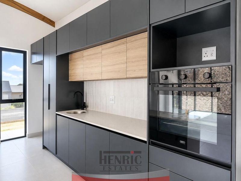 Modern kitchen featuring sleek dark cabinetry, light wood accents, stone countertops, built-in oven, and a minimalist tiled backsplash with a view to the exterior.
