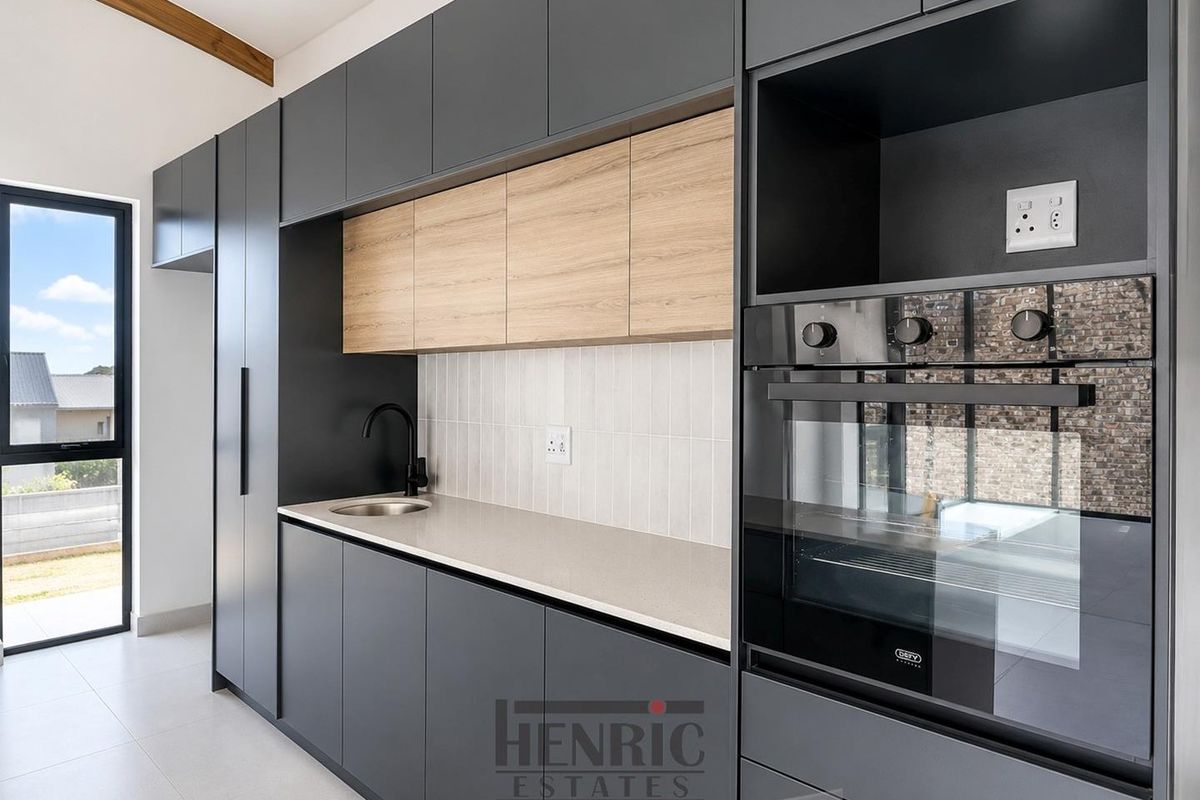 Modern kitchen featuring sleek dark cabinetry, light wood accents, stone countertops, built-in oven, and a minimalist tiled backsplash with a view to the exterior.