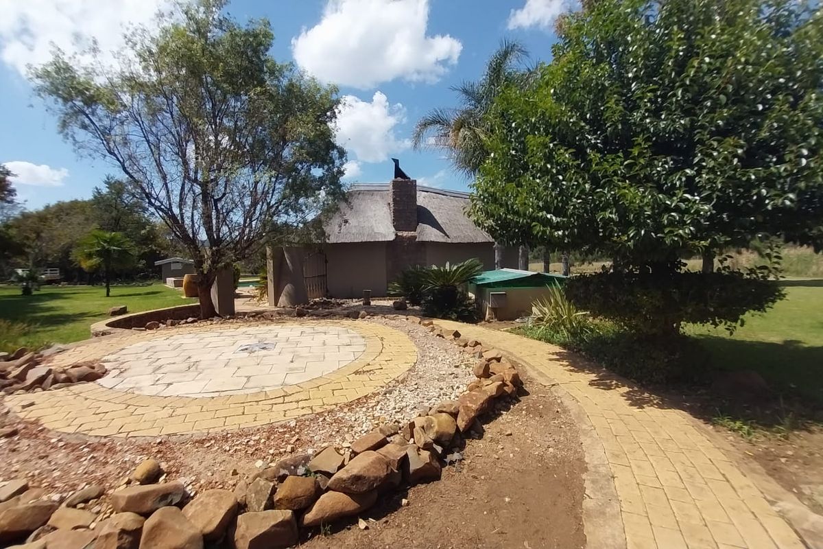 A rear view of a property featuring a thatched roof structure, a circular paved patio area, and a landscaped garden with mature trees.