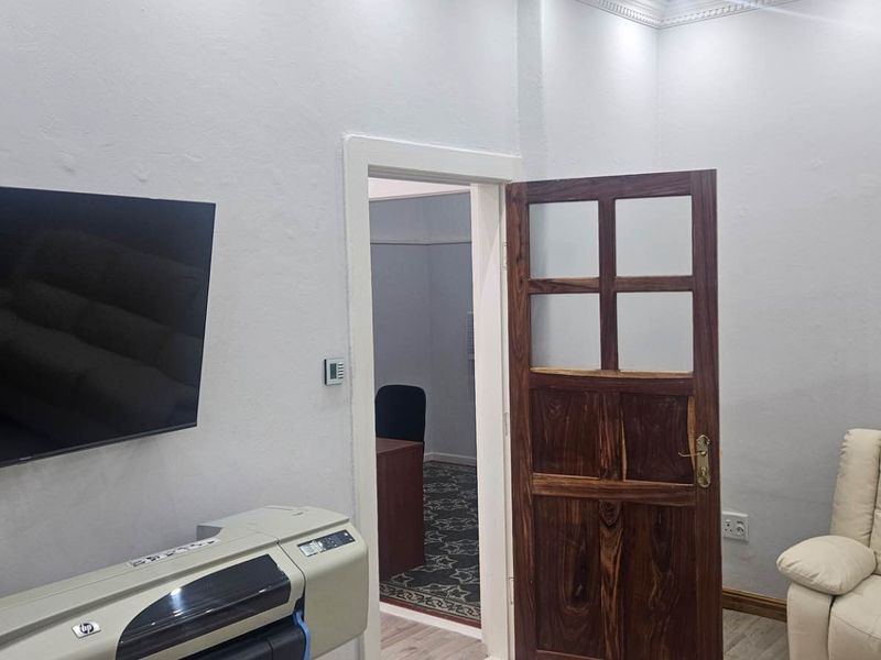 A functional office space featuring a wooden door, neutral wall finishes, and a view into an adjacent workspace.