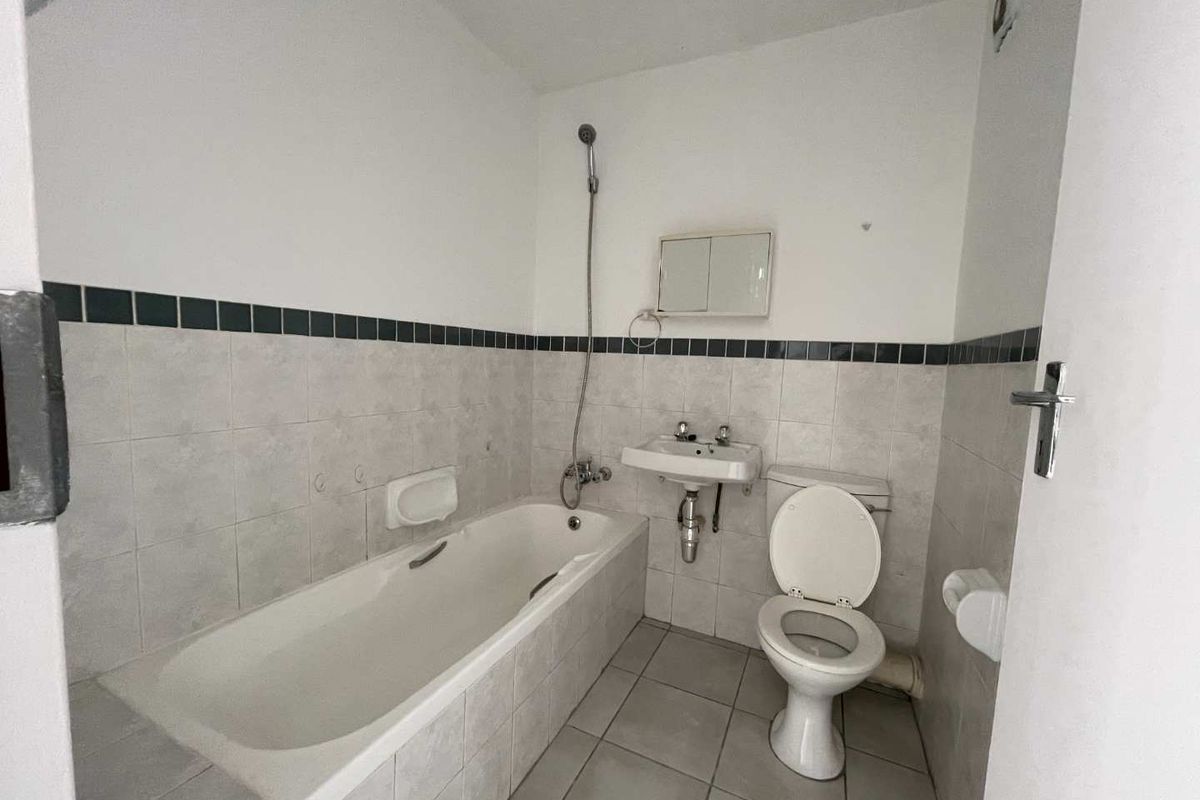 A functional bathroom featuring a bathtub with a handheld shower, a wall-mounted basin, and a toilet, finished with neutral wall and floor tiling.