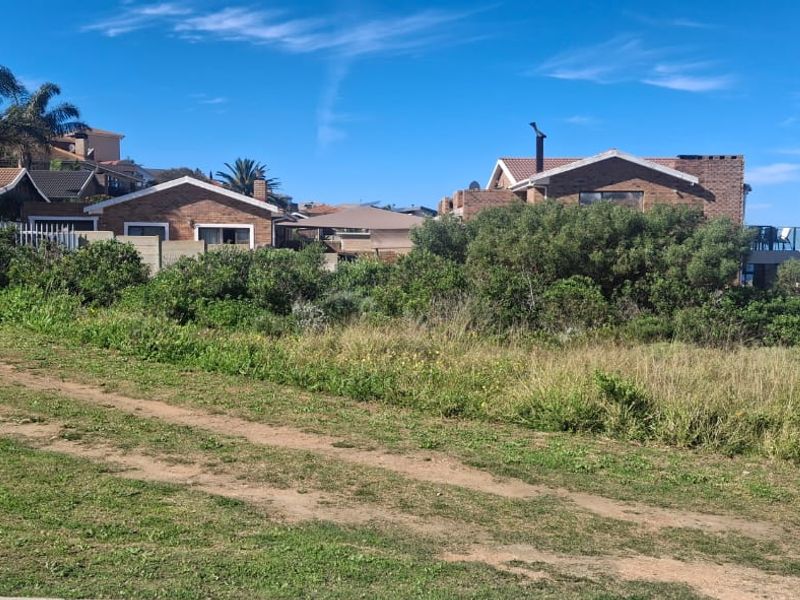 A view of a vacant residential plot featuring an unpaved access track, natural coastal vegetation, and surrounding established residential properties.