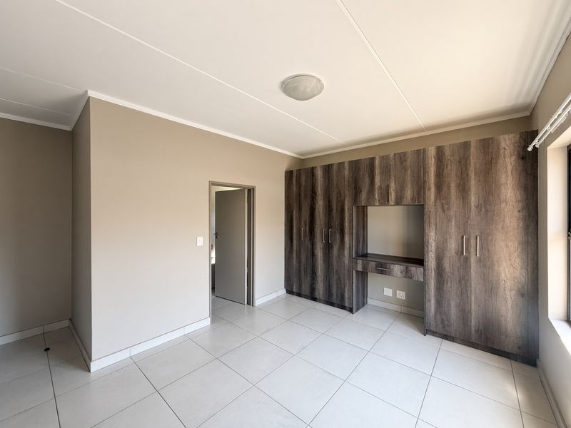 A spacious, empty bedroom featuring neutral wall tones, large-format floor tiles, and extensive dark wood-grain built-in cupboards with an integrated desk area.