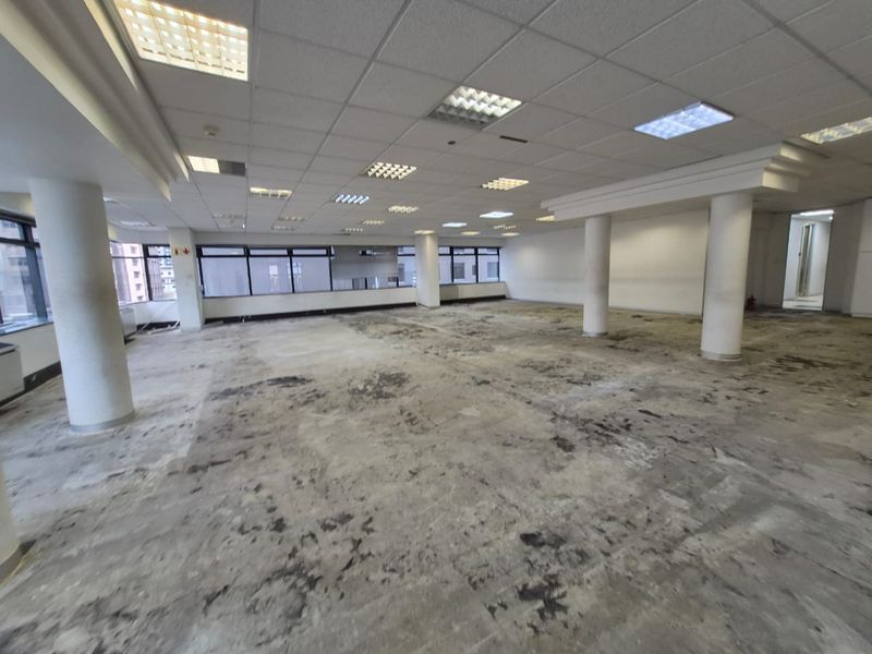 A large, open-plan office space featuring a suspended ceiling, recessed lighting, and structural columns, currently requiring significant flooring repairs and cosmetic updates.
