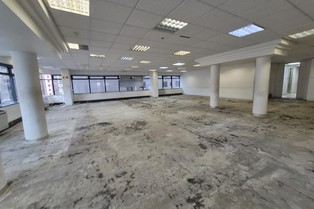 A large, open-plan office space featuring a suspended ceiling, recessed lighting, and structural columns, currently requiring significant flooring repairs and cosmetic updates.