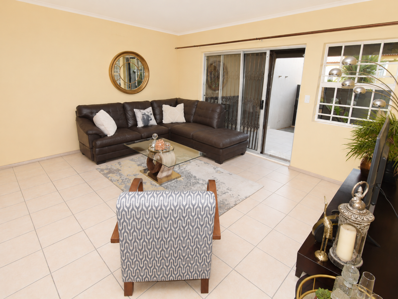 A spacious living room featuring neutral tiled flooring, large sliding glass doors with security gates, and natural light through a window with burglar bars.