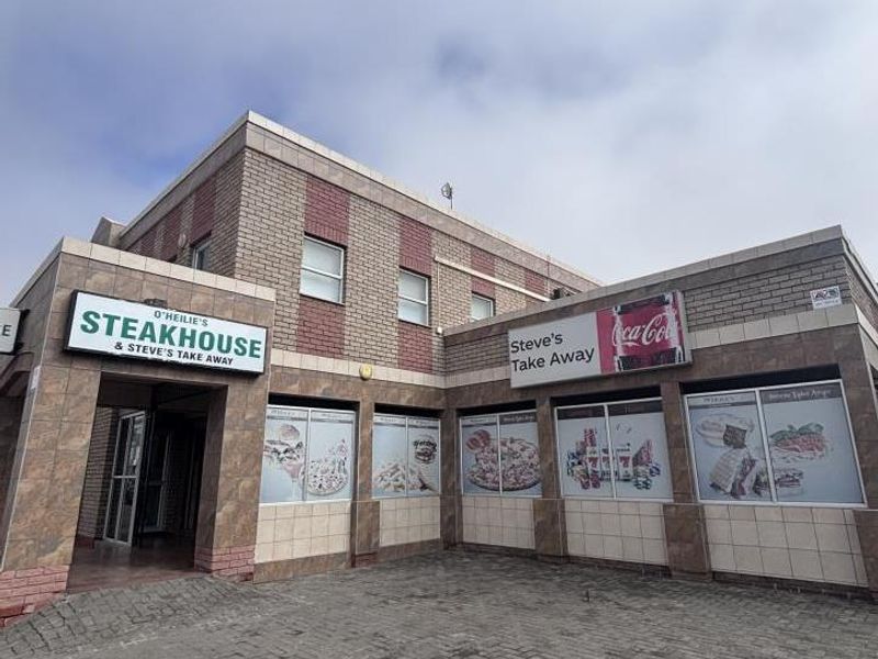 Front exterior view of a mixed-use commercial building featuring brick and tiled facade, paved driveway, and prominent signage for a steakhouse and takeaway.