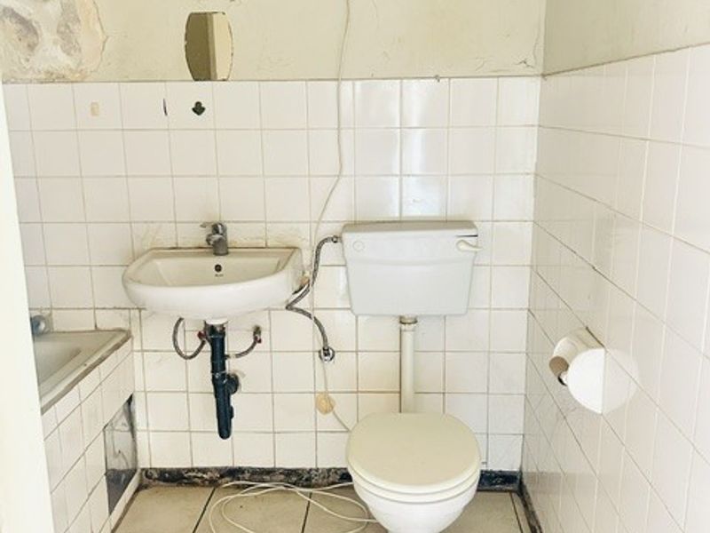 A dated bathroom requiring significant renovation, featuring a wall-mounted basin, toilet, and tiled finishes with visible signs of wear and structural deterioration.