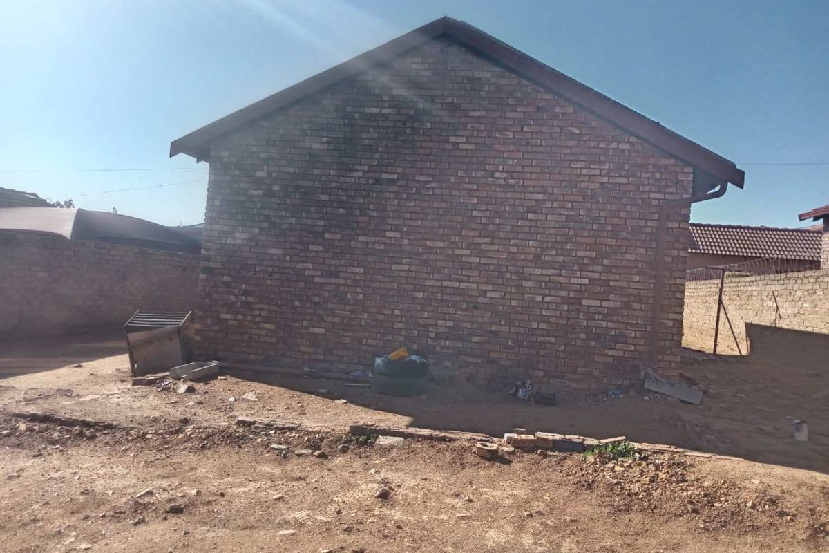 A view of the side exterior brick facade of the house, featuring a gabled roofline and surrounding unpaved yard area.