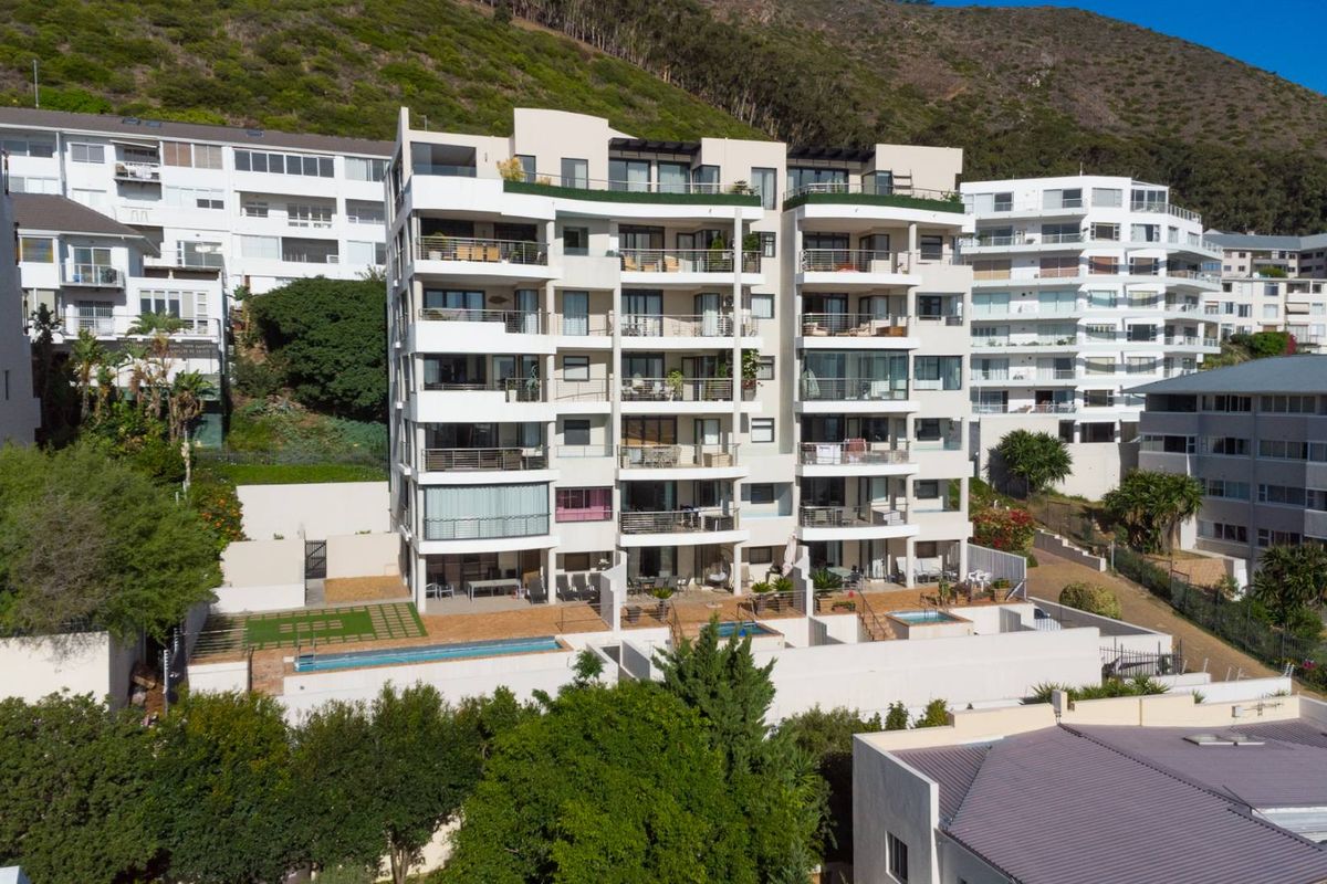Aerial view of a multi-story apartment complex featuring private balconies, ground-floor terraces, and swimming pools set against a mountain backdrop.