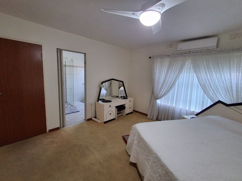 A spacious bedroom featuring carpeted flooring, a ceiling fan, a wall-mounted air conditioning unit, and direct access to an en-suite bathroom.