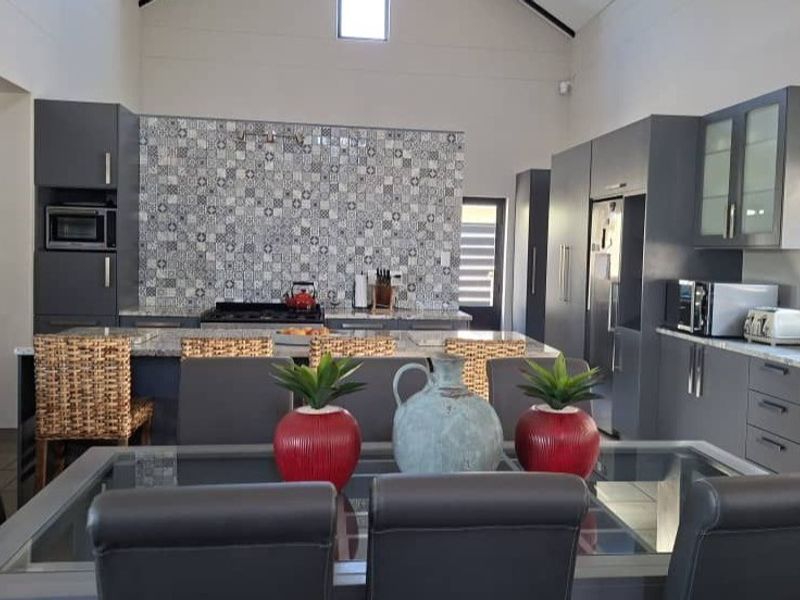 Spacious open-plan kitchen and dining area featuring modern grey cabinetry, stone countertops, a striking patterned tile backsplash, and a high vaulted ceiling.