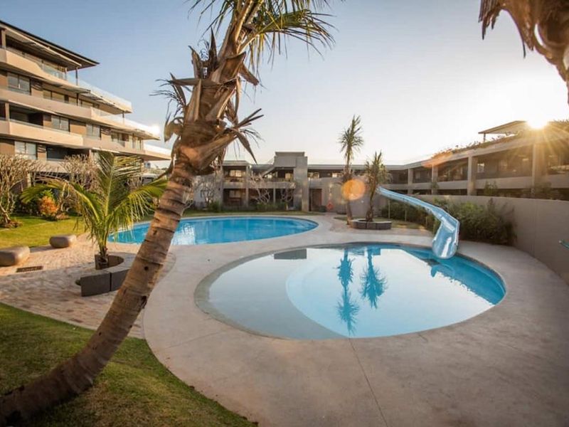 A well-maintained communal pool area featuring two circular swimming pools, a water slide, and landscaped garden surrounds within an apartment complex.