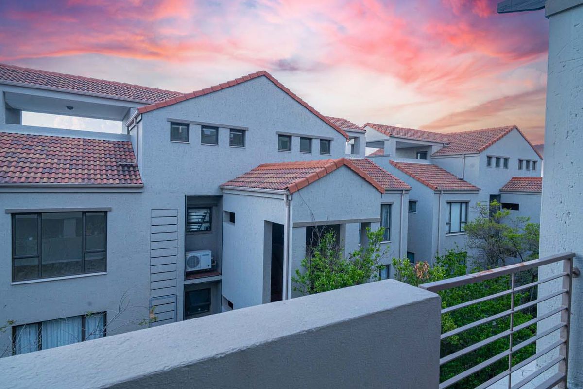 View of the apartment complex exterior featuring terracotta roof tiles, stucco facade, and a metal balustrade balcony at sunset.