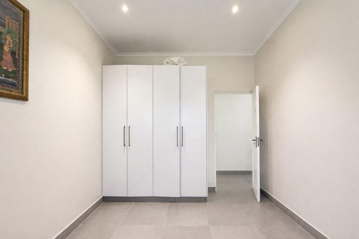 A clean, neutral bedroom featuring built-in white cabinetry, recessed ceiling lighting, and modern tiled flooring, leading to an adjacent room.