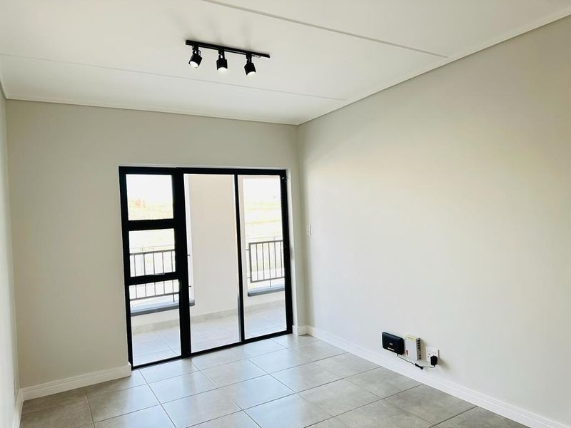 A bright, empty living room featuring neutral tiled floors, modern track lighting, and a sliding glass door leading to an outdoor balcony.