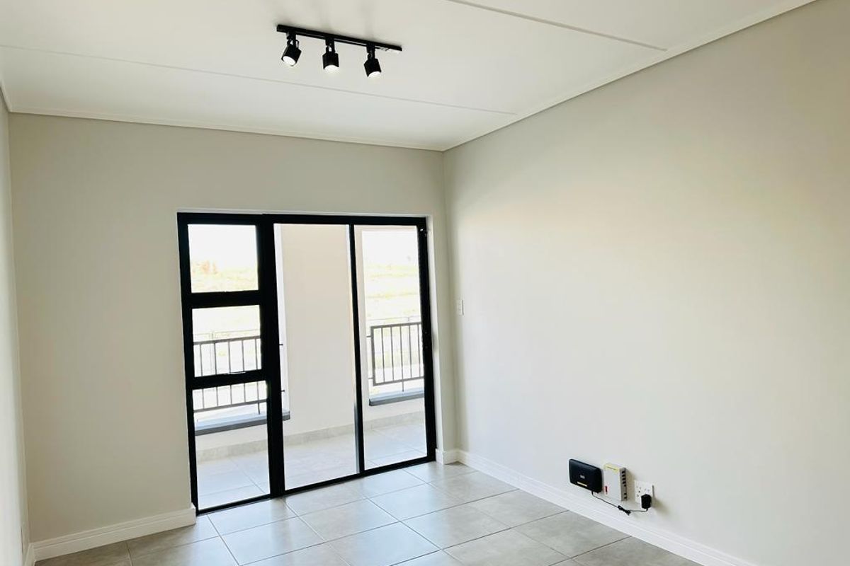 A bright, empty living room featuring neutral tiled floors, modern track lighting, and a sliding glass door leading to an outdoor balcony.