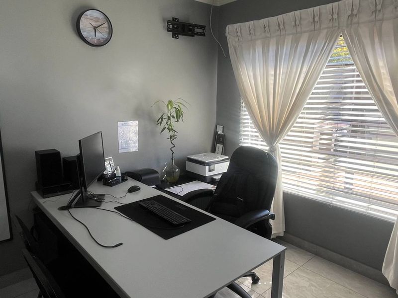 A functional home office space featuring tiled flooring, neutral wall tones, and window treatments including venetian blinds and curtains.