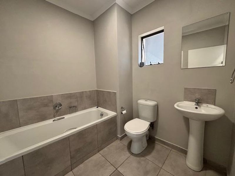 A clean and functional bathroom featuring a bathtub, pedestal sink, toilet, and neutral wall tiles, offering a simple and well-maintained space.