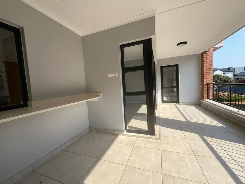 A spacious, covered balcony featuring neutral tiled flooring, modern black aluminium framed glass doors, and a built-in shelf, offering a clean outdoor living space.