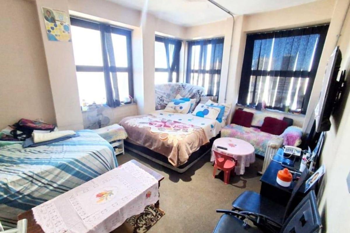A bedroom featuring large windows providing natural light, with visible exposed electrical conduit and dated flooring finishes.