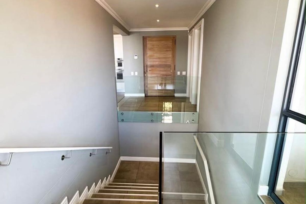 A bright, modern hallway featuring a tiled staircase with a sleek glass balustrade, crown molding, and a solid timber entrance door.