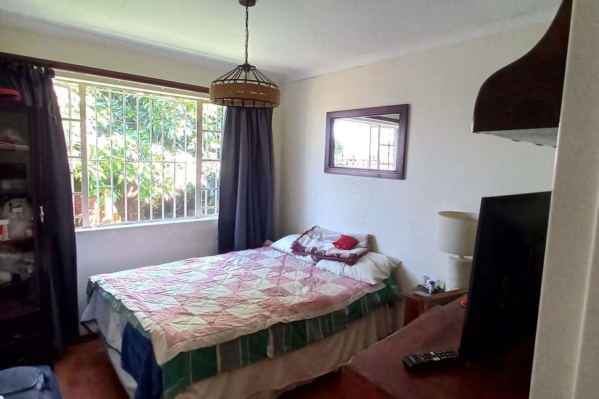 A functional bedroom featuring a large window with security bars, providing natural light and a view of the garden area.