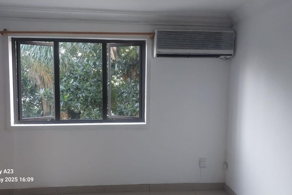 An empty bedroom featuring tiled flooring, a wall-mounted air conditioning unit, and a casement window overlooking greenery.