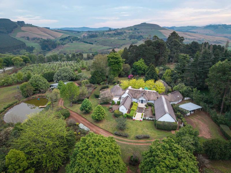 Aerial view of a sprawling farm property featuring a traditional thatched-roof residence, landscaped gardens, a private pond, and surrounding rural landscape.