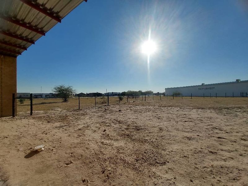 An expansive, unpaved industrial yard area featuring a corrugated metal roof overhang and perimeter wire fencing under a clear sky.