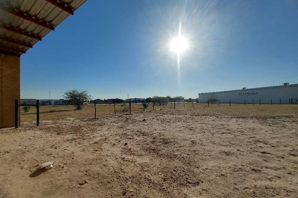 An expansive, unpaved industrial yard area featuring a corrugated metal roof overhang and perimeter wire fencing under a clear sky.