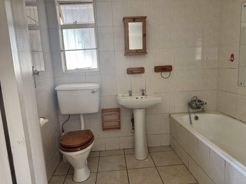 A dated bathroom featuring a pedestal basin, standard toilet, and bathtub with wall-to-wall tiling and a small window for natural light.