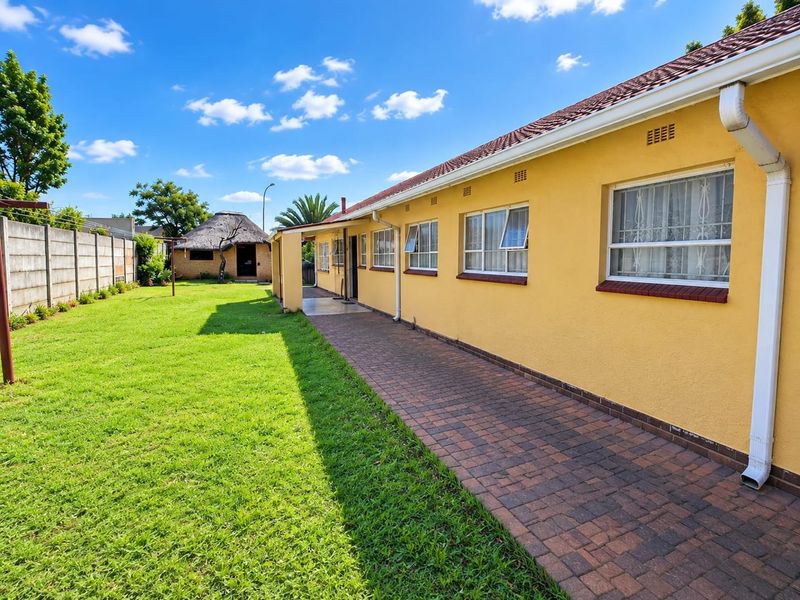 Rear view of the property featuring a paved walkway, well-maintained lawn, precast boundary wall, and a detached thatched lapa in the garden.