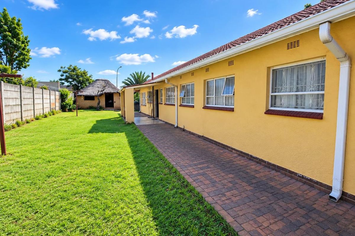 Rear view of the property featuring a paved walkway, well-maintained lawn, precast boundary wall, and a detached thatched lapa in the garden.