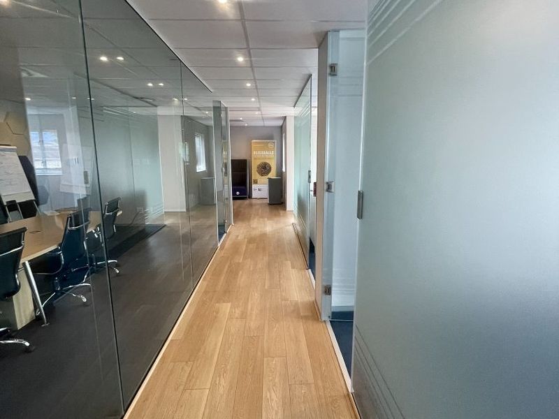 A modern office hallway featuring wood-look flooring, suspended ceilings with recessed lighting, and glass partition walls creating a bright, professional workspace environment.
