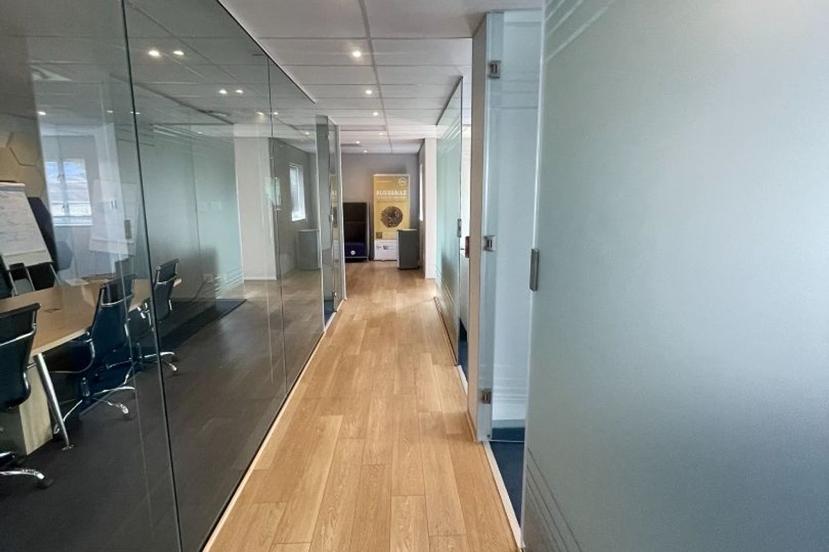 A modern office hallway featuring wood-look flooring, suspended ceilings with recessed lighting, and glass partition walls creating a bright, professional workspace environment.