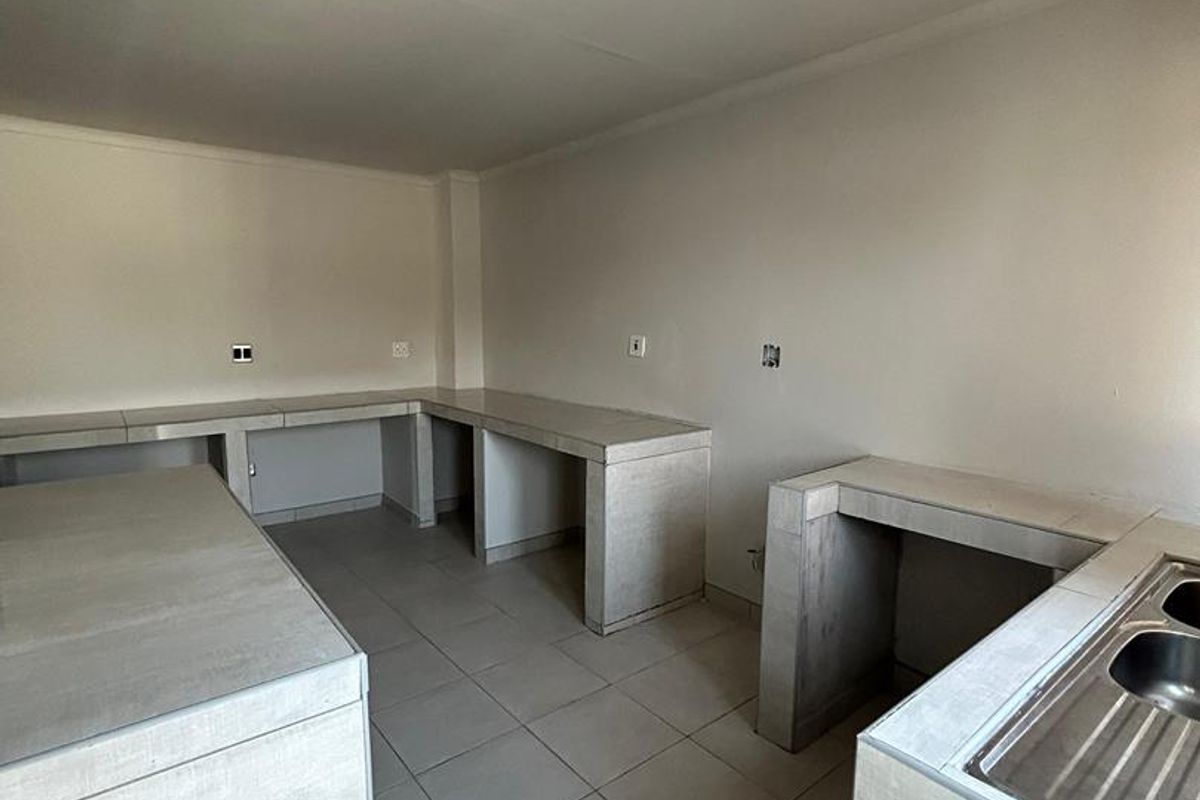 A minimalist kitchen featuring concrete-style countertops, a stainless steel sink, and neutral tiled flooring in an open, empty layout.