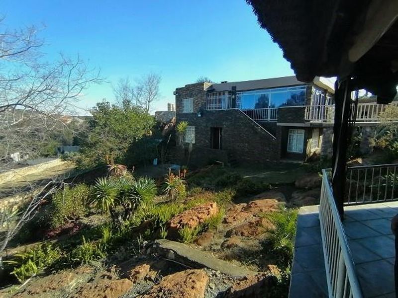 Rear view of a multi-level stone-clad house featuring a tiled patio, metal balustrades, and expansive windows overlooking a landscaped rocky garden.