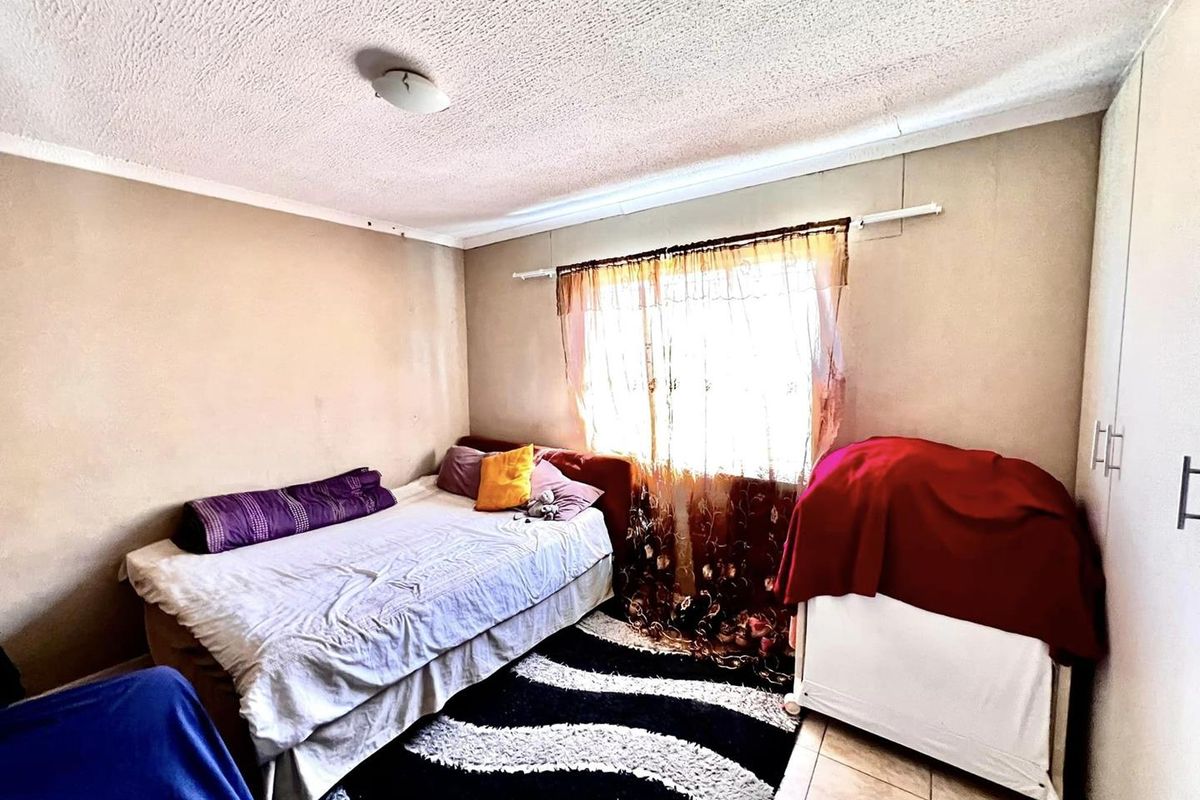 A bedroom featuring tiled flooring, built-in cupboards, and a window with curtain rails, offering a functional space in need of cosmetic updates.