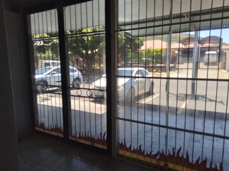Interior view of a retail entrance featuring large glass storefront windows with security gates and tiled flooring, looking out onto a parking area.