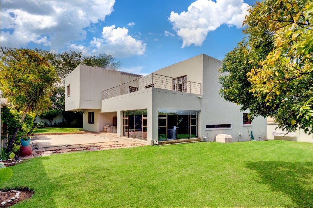 Rear view of a modern two-story house featuring a spacious lawn, paved patio area, and an upper-level balcony with metal railings.