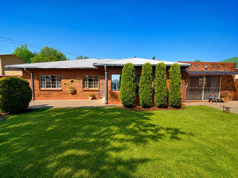 A well-maintained rear exterior view of a brick house featuring a corrugated metal roof, security bars on windows, a paved patio, and a manicured lawn.