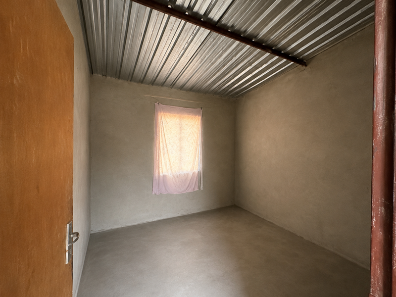 A simple, unfinished bedroom featuring plastered walls, a cement screed floor, and a corrugated metal ceiling.